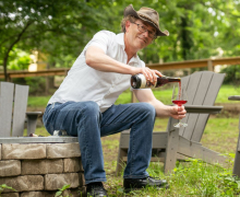 Erik Martella Erik Martella sits on a stone wall and smiles while pouring himself a glass of red wine.