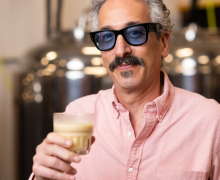 A gray-haired man with a mustache, dark glasses, and a pink shirt holds a beige beverage in a fermentation sciences laboratory.