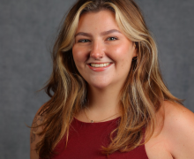 Smiling woman with long wavy blonde-brown hair, nose ring, and burgundy sleeveless top, posed against a gray studio background